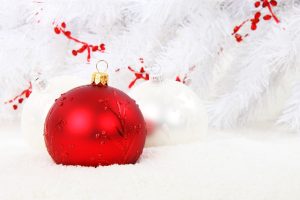 two Christmas ornaments, one red, one white, on snow in front of a white tree with red holly